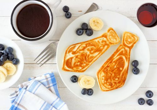 A breakfast plate with two fish-shaped pancakes, bananas, blueberries, a cup of coffee, and a drink, alongside a blue-checkered napkin and utensils.