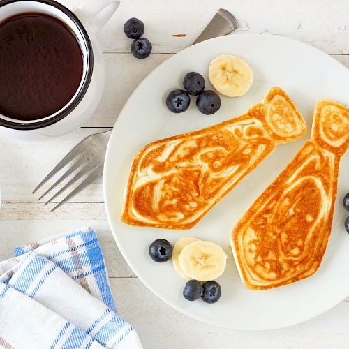 A breakfast plate with two fish-shaped pancakes, bananas, blueberries, a cup of coffee, and a drink, alongside a blue-checkered napkin and utensils.