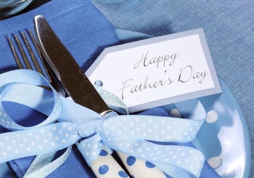 A table setting with a "Happy Father's Day" card, blue theme, cutlery tied with polka dot ribbons on a plate and napkin, and a glass of water.