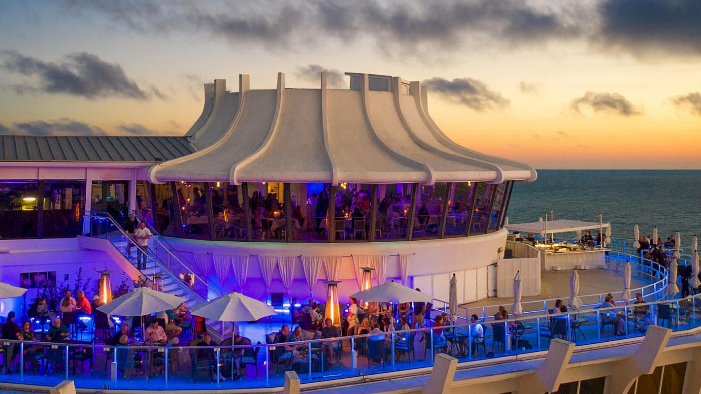 An outdoor dining area, likely on a ship, illuminated with blue lighting and white umbrellas, set against a scenic sunset over the ocean.