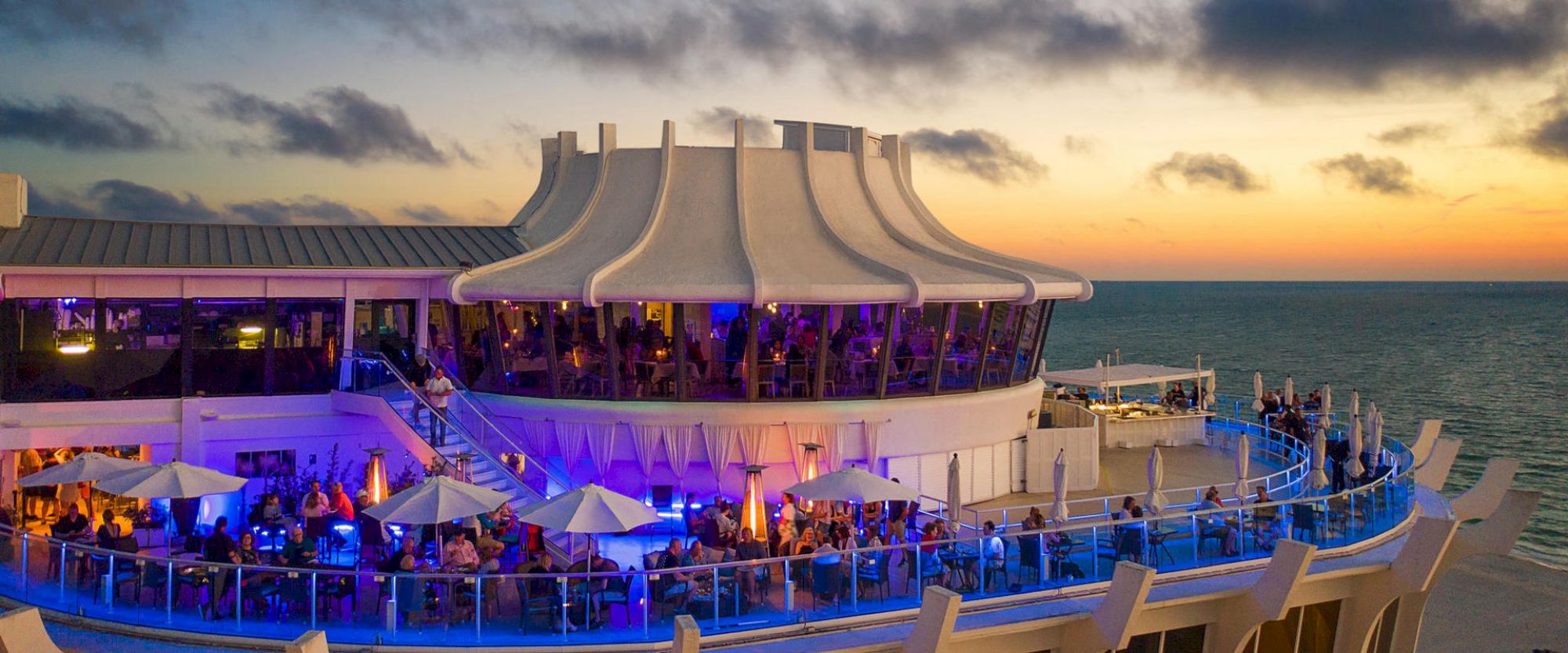 An outdoor dining area, likely on a ship, illuminated with blue lighting and white umbrellas, set against a scenic sunset over the ocean.