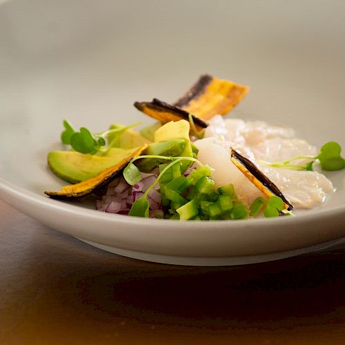 A gourmet dish features sliced avocado, microgreens, chopped onions, and fish or seafood in a white bowl, garnished with plantain chips.