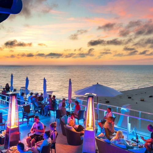 A vibrant beachside rooftop bar at sunset, with patrons enjoying drinks and the ocean view under colorful ambient lighting.