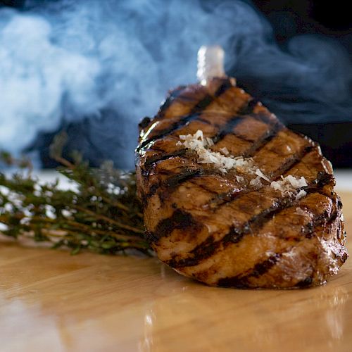 A grilled steak garnished with herbs and salt, presented on a wooden board with smoke rising in the background.