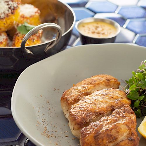 A plate of roasted chicken, a lemon wedge, and microgreens, with a side bowl of breaded snacks and a dipping sauce on a blue-tiled surface.