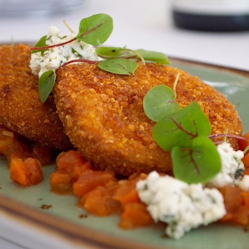 The image shows a plate of crispy golden brown patties garnished with fresh greens and crumbled cheese, served on a bed of diced tomatoes.