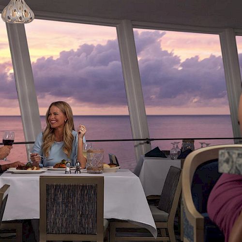 Three people are at a restaurant with large windows and an ocean view at sunset. A couple dines while a woman drinks alone at a nearby table.