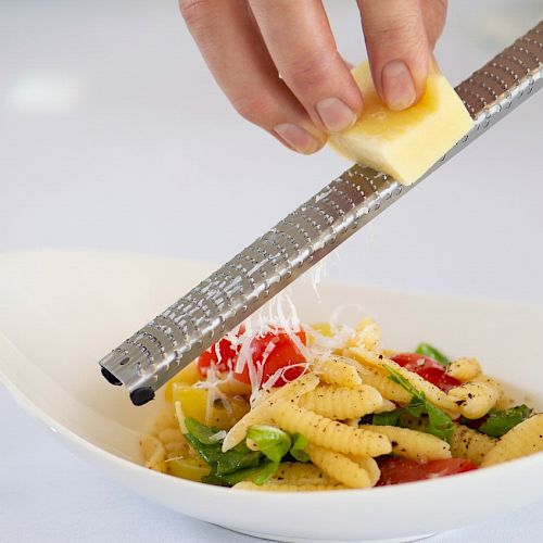 A hand is grating cheese over a bowl of pasta with vegetables and sauce, using a fine grater.