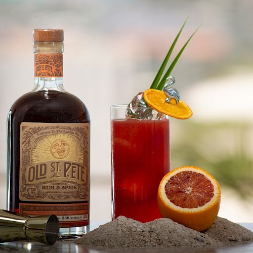 A bottle of Old St. Pete Rum & Spice, a cocktail garnished with fruit and leaves, a citrus half, a jigger, and some sand are on the table.