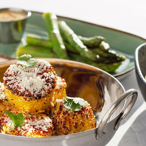 The image shows a plated meal with corn on the cob topped with cheese, asparagus spears, and a side of mashed potatoes garnished with chives.