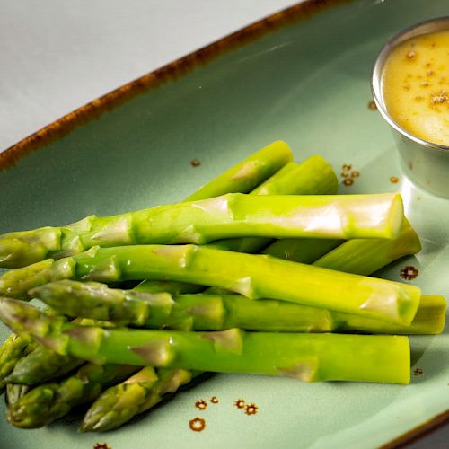 The image shows a plate of asparagus spears served with a small metal cup of dipping sauce on the side.