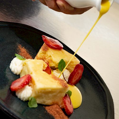 The image shows a dessert plate with pieces of cake, strawberries, cream, and sauce being poured over the dish, garnished with greens and spices.