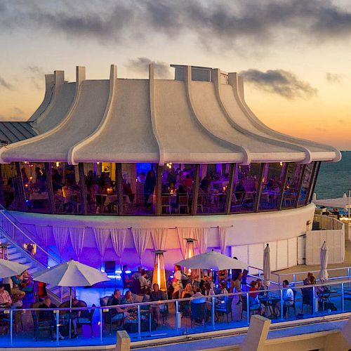 The image shows a rooftop restaurant or bar with patrons under umbrellas at sunset, set against an ocean backdrop.