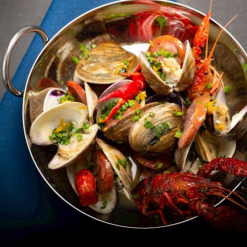 The image shows a dish of assorted shellfish, including clams and crawfish, garnished with herbs and served in a metal bowl with handles.