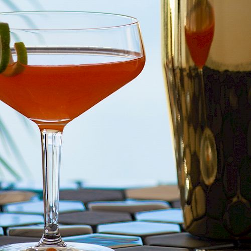 A cocktail with a lime twist in a coupe glass beside a shiny cocktail shaker on a patterned surface.