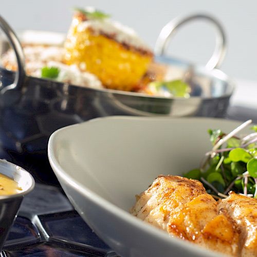 The image shows a plated dish with grilled fish, greens, and a small bowl of sauce. In the background, there is a pot with some additional food.