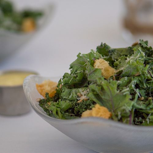 A fresh green salad with croutons, served in a glass bowl, with a side of dressing in a small metal cup on a white tablecloth.