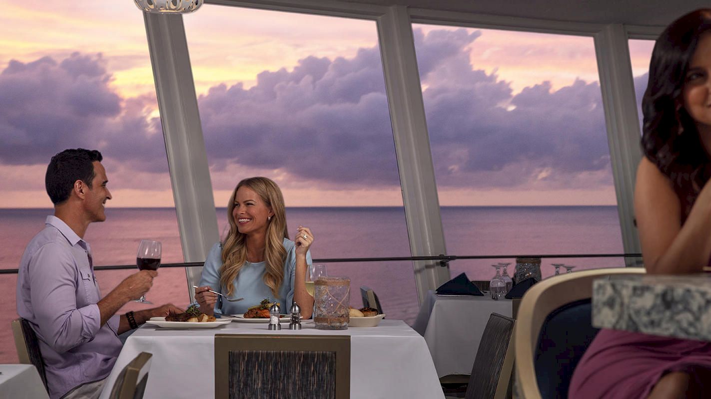 Three people in a restaurant with an ocean view at sunset. A couple is dining and smiling while another woman enjoys a drink alone at the bar.