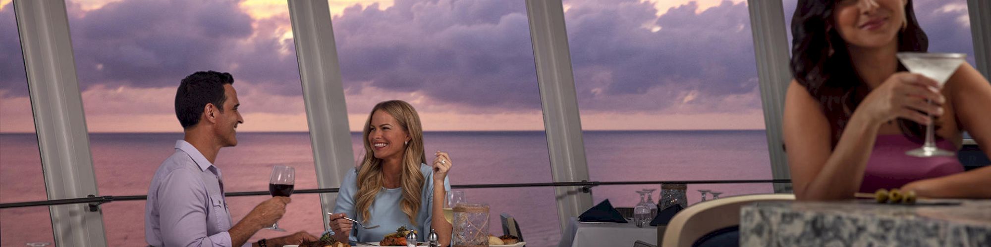 Three people in a restaurant with an ocean view at sunset. A couple is dining and smiling while another woman enjoys a drink alone at the bar.