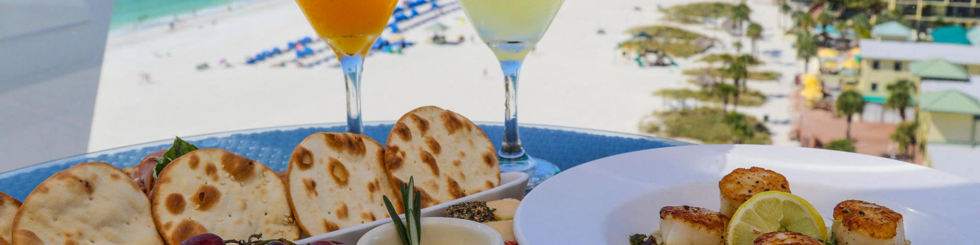 A seaside dining setup featuring cocktails, a platter with bread, olives, grapes, and a dish of grilled scallops with lemon on a beach backdrop.