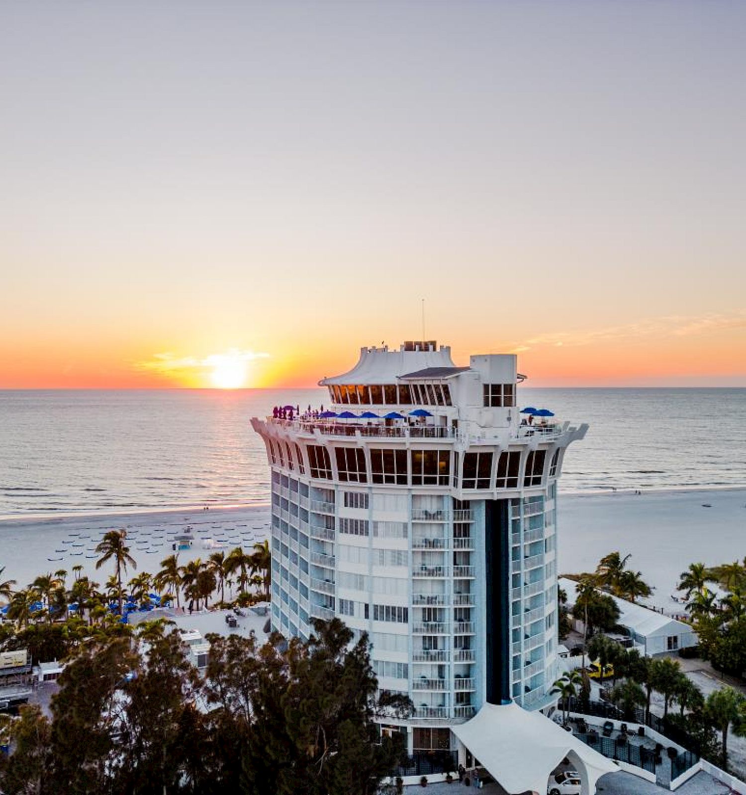 A beachfront hotel with a unique round structure, surrounded by palm trees, overlooking the ocean at sunset.