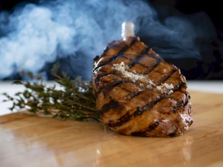 A succulent grilled steak with crisscross marks, garnished with herbs and sprinkled salt, resting on a wooden cutting board with steam rising.