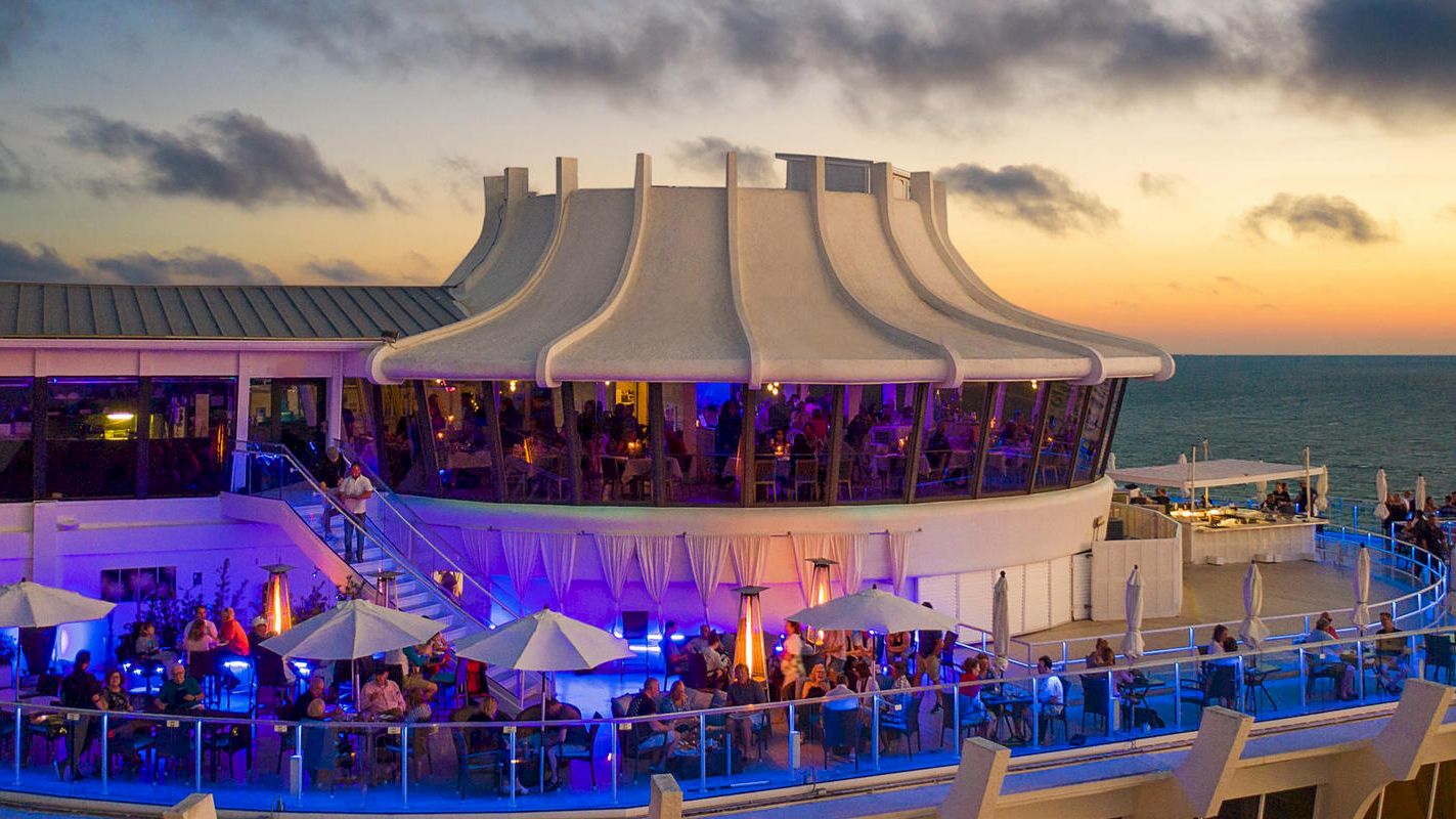 The image shows a vibrant, multi-level restaurant with outdoor seating on a deck overlooking the ocean at sunset.