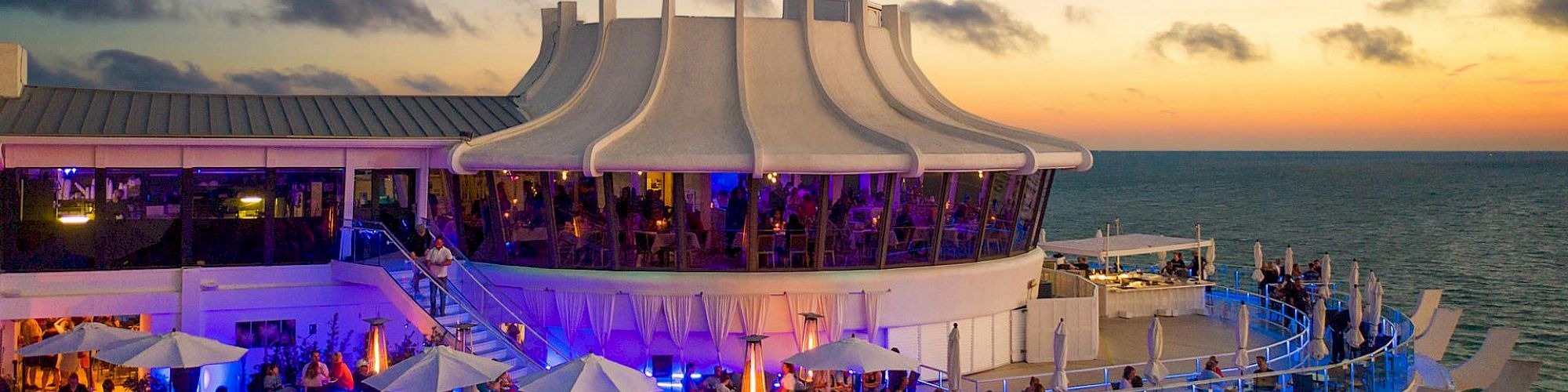 An outdoor restaurant or bar with people dining under umbrellas, located on a curved deck overlooking the ocean at sunset.