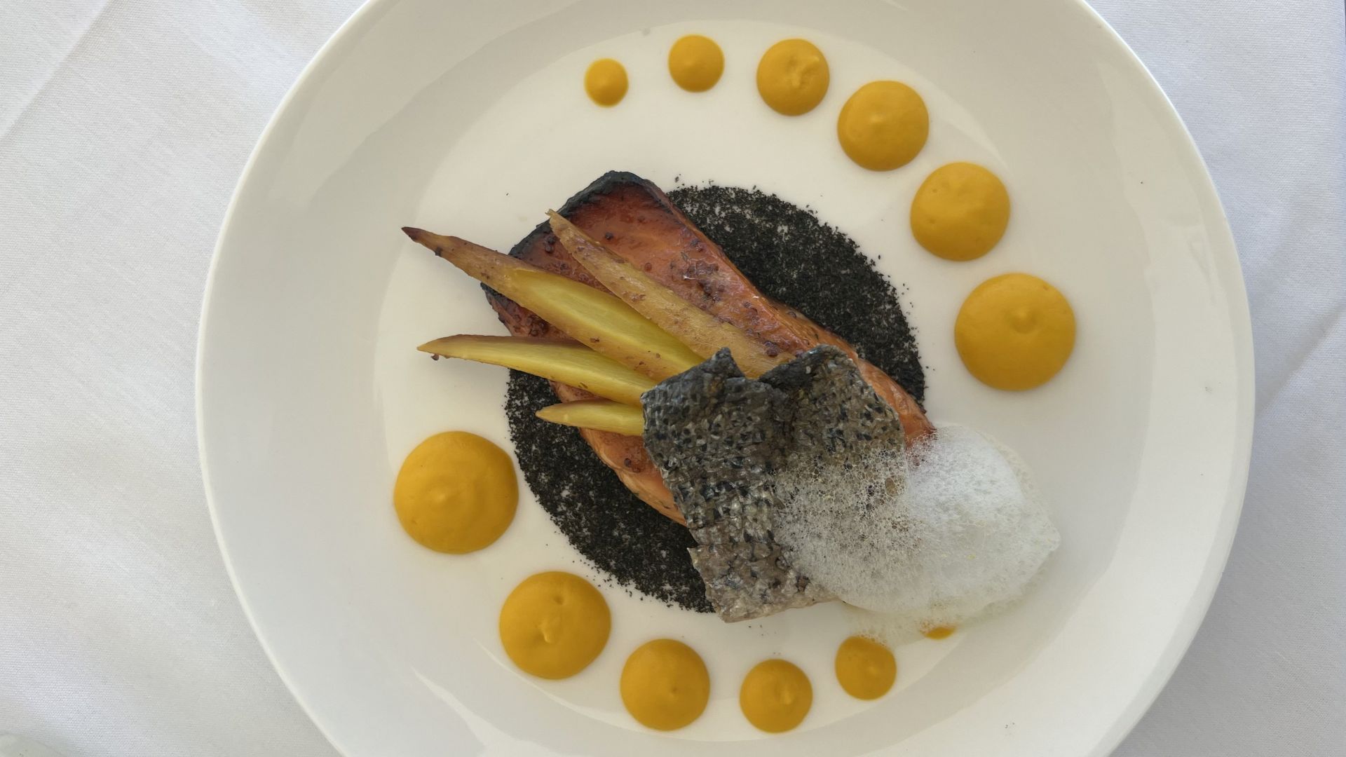 A plated gourmet dish featuring fish, foam, and vegetable garnishes, surrounded by dollops of a yellow sauce on a white plate.