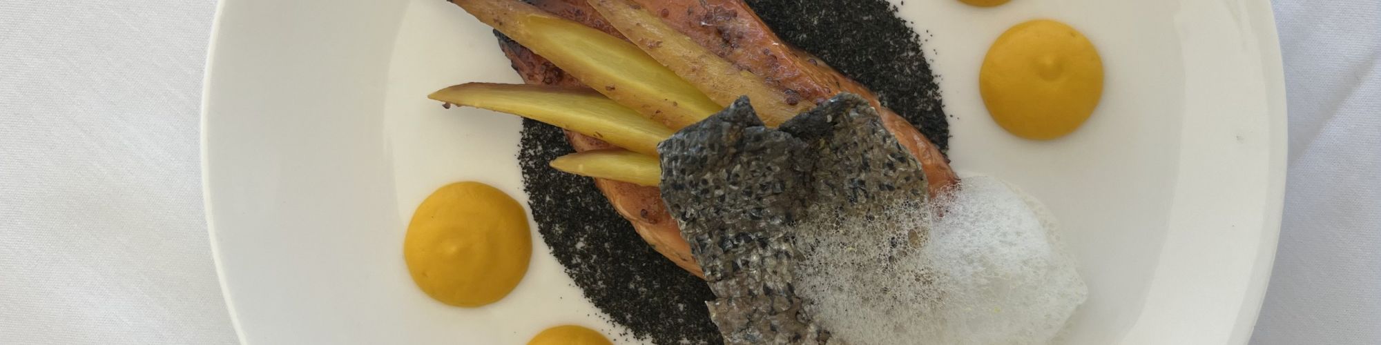 A plated gourmet dish featuring fish, foam, and vegetable garnishes, surrounded by dollops of a yellow sauce on a white plate.