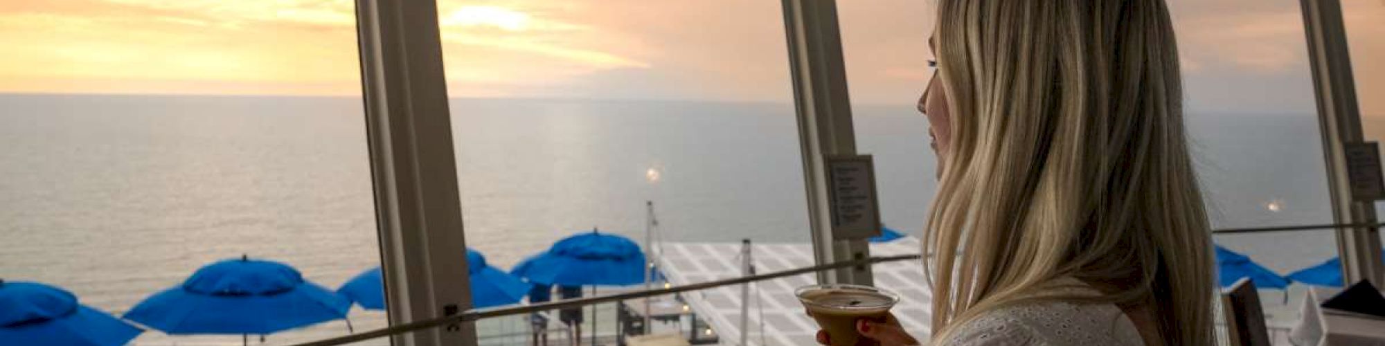 A person is indoors enjoying a drink while looking at the ocean view through large windows with blue umbrellas outside.