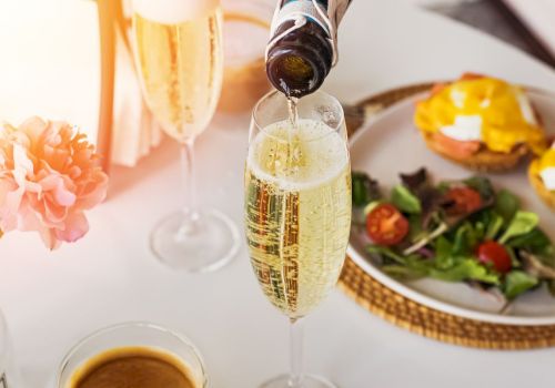 A bottle pours champagne into a flute on a table with a plate of eggs Benedict, salad, a flower vase, and a cup of coffee.