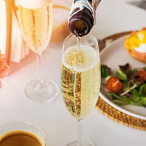 A bottle pours champagne into a flute on a table with a plate of eggs Benedict, salad, a flower vase, and a cup of coffee.