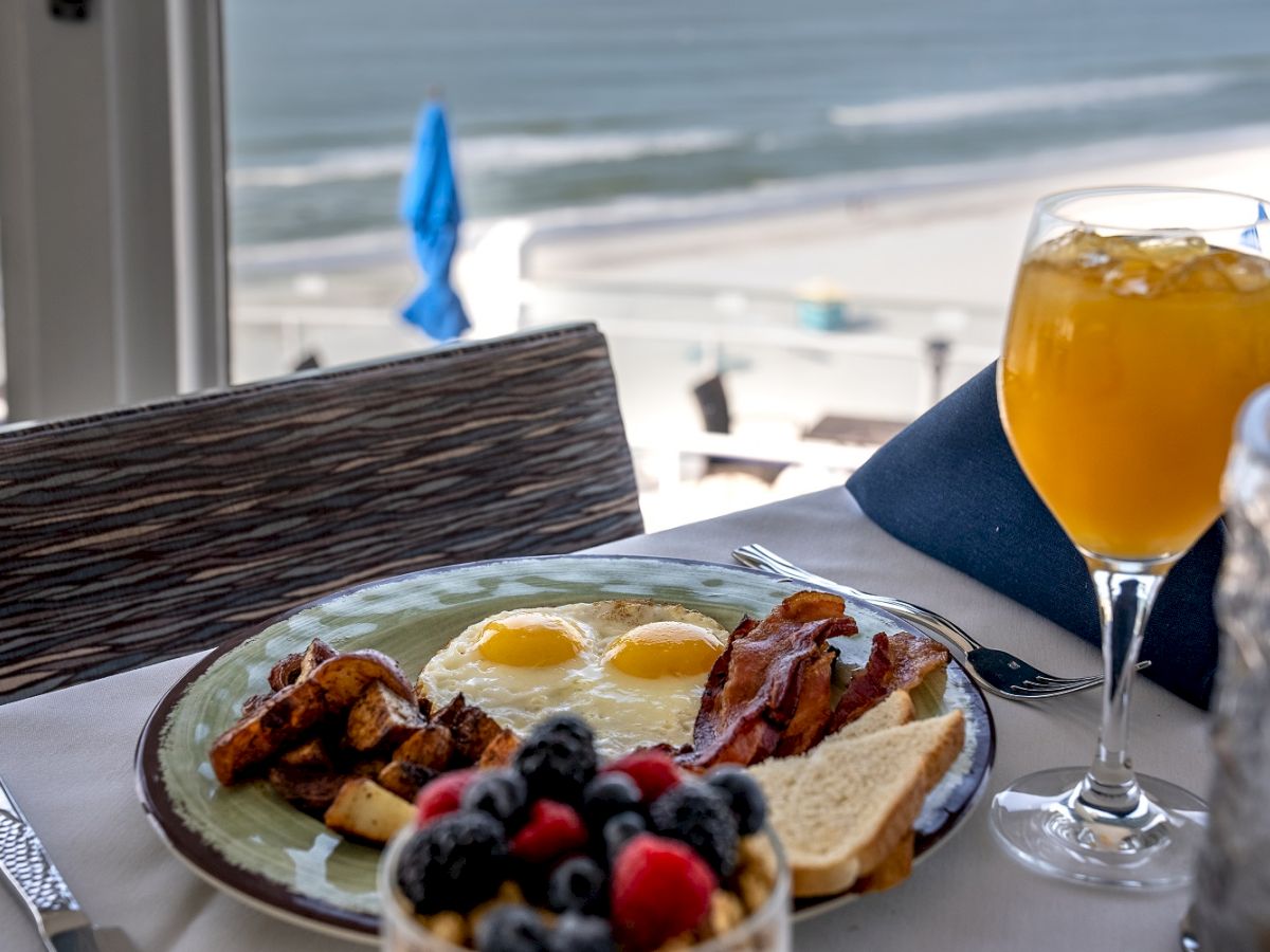 A breakfast table by the beach with a plate of eggs, bacon, toast, a bowl of berries, and a glass of orange juice.