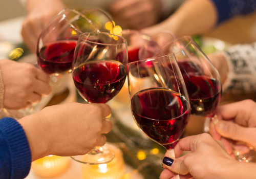 A group of people clinking glasses of red wine together in a celebratory toast over a table with candles and food, creating a festive atmosphere.