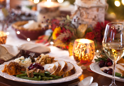 A festive dinner table with plates of food, candles, and wine glasses set against a background of warm lights and holiday decorations.