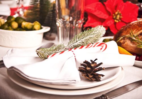 A festive holiday table setting featuring white plates, napkins with a candy cane and pinecone, and a centerpiece with poinsettia and food.