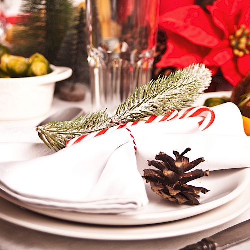 A festive holiday table setting featuring white plates, napkins with a candy cane and pinecone, and a centerpiece with poinsettia and food.