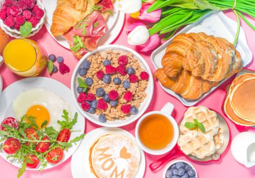 The image features a breakfast spread with croissants, cereal with berries, pancakes, waffles, a fried egg, cherry tomatoes, juice, coffee, and flowers.