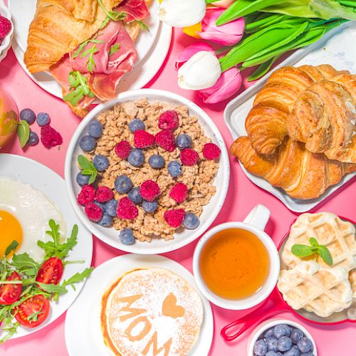 The image features a breakfast spread with croissants, cereal with berries, pancakes, waffles, a fried egg, cherry tomatoes, juice, coffee, and flowers.