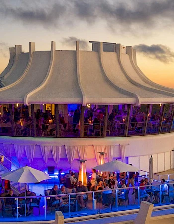 A vibrant outdoor dining area with patrons under white umbrellas, situated on a platform overlooking the ocean at sunset, featuring modern architecture.